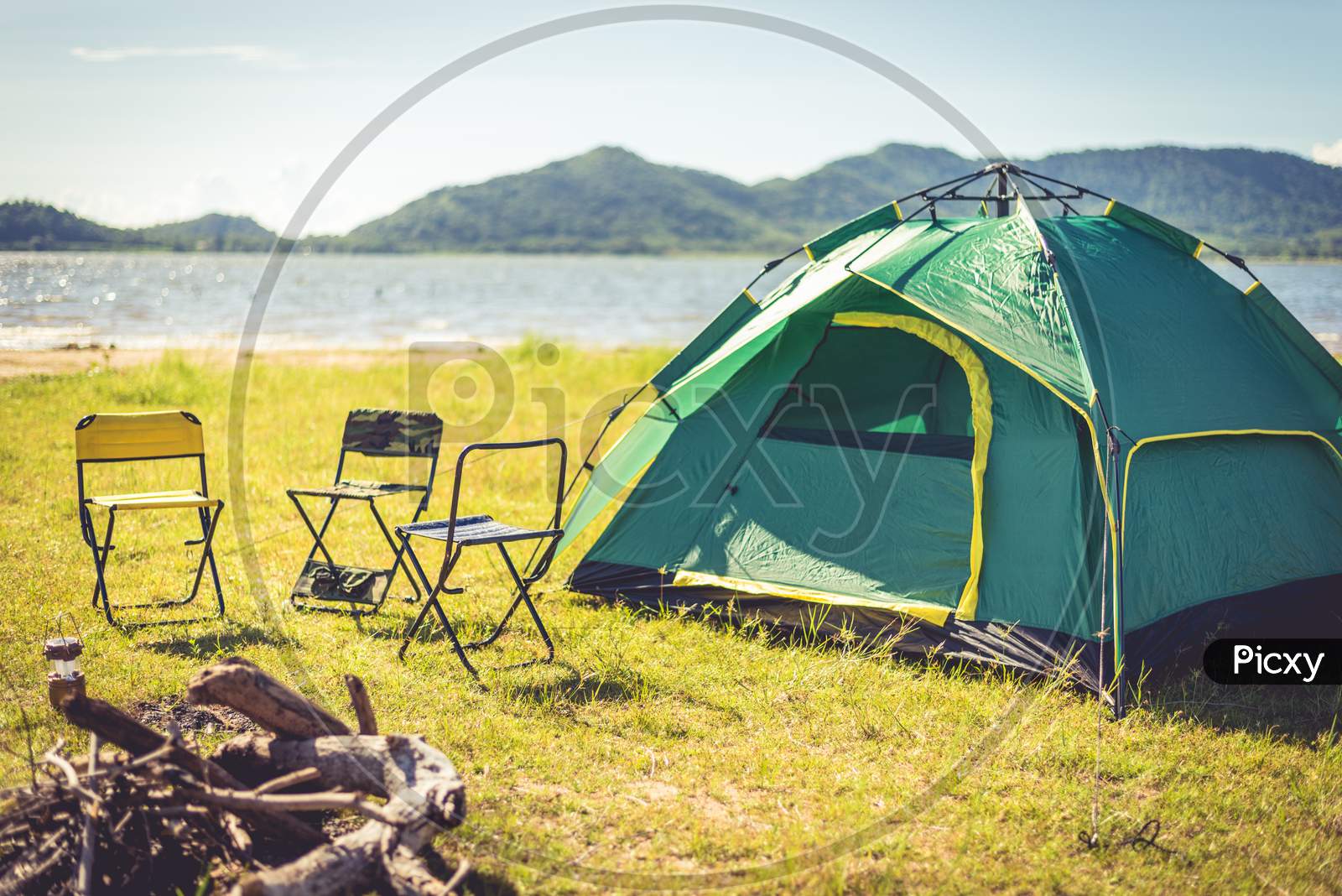 Camping Tent With Extinguished Bonfire In The Green Field Meadow, Lake And Mountain Background. Picnic And Travel Concept. Nature Theme.