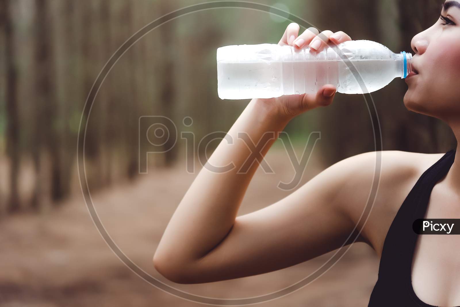 Close Up Of Beauty Asian Woman  Drinking Purify Water By Right Hand After Exercise In Forest. Thirsty Girl In Black Sport Wear. Workout And Sports Concept. People And Outdoor Nature Theme.