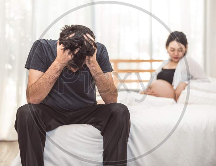 Worried Stress Man Sitting On Bed With Hand On Forehead In Bedroom In Serious Mood Emotion With Pregnant Wife Woman Background. Major Depressive Disorder Called Mdd Concept. Physical Healthcare