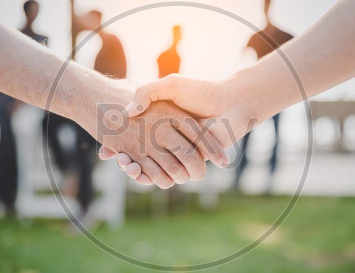 Handshake Of Two Business People At Outdoors. Business And Nature Concept. Meeting And Partnership Theme. Blurry People In Background Elements.