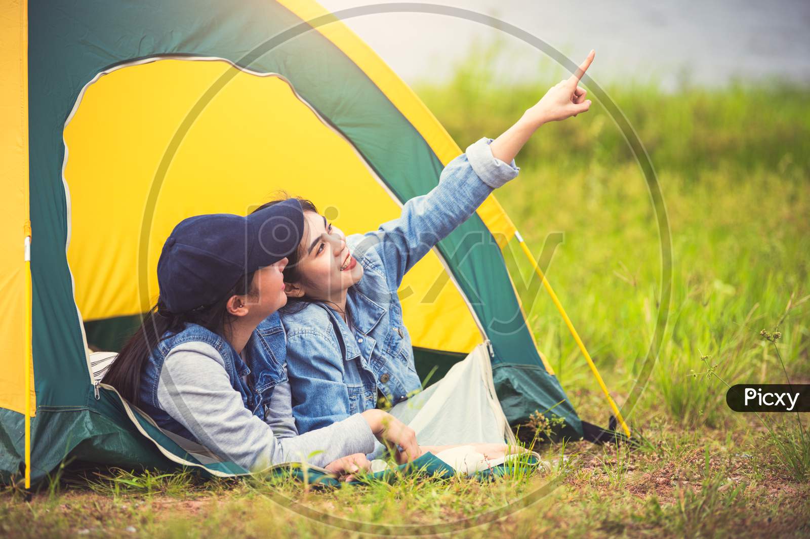 Two Close Friend Asian Friendship Relax In Camping Tent In Green Meadow On Lake Side View Background. Girl Pointing Finger To Sky. People Lifestyle Travel On Vacation Concept. Summer Picnic Activity
