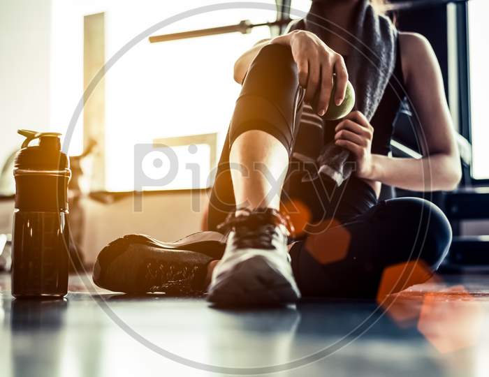 Sport Woman Sitting And Resting After Workout Or Exercise In Fitness Gym With Protein Shake Or Drinking Water On Floor. Relax Concept. Strength Training And Body Build Up Theme. Warm And Cool Tone
