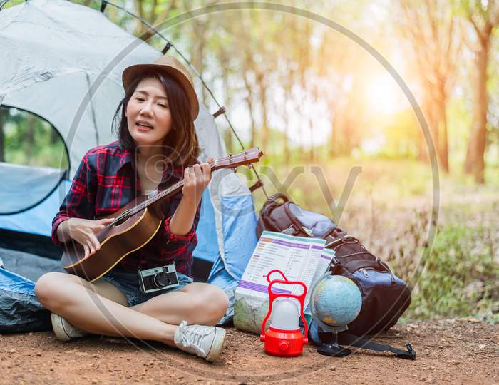 Beautiful Asian Woman Playing Ukulele In Front Of Camping Tent In Pine Woods. People And Lifestyles Concept. Adventure And Travel Theme.