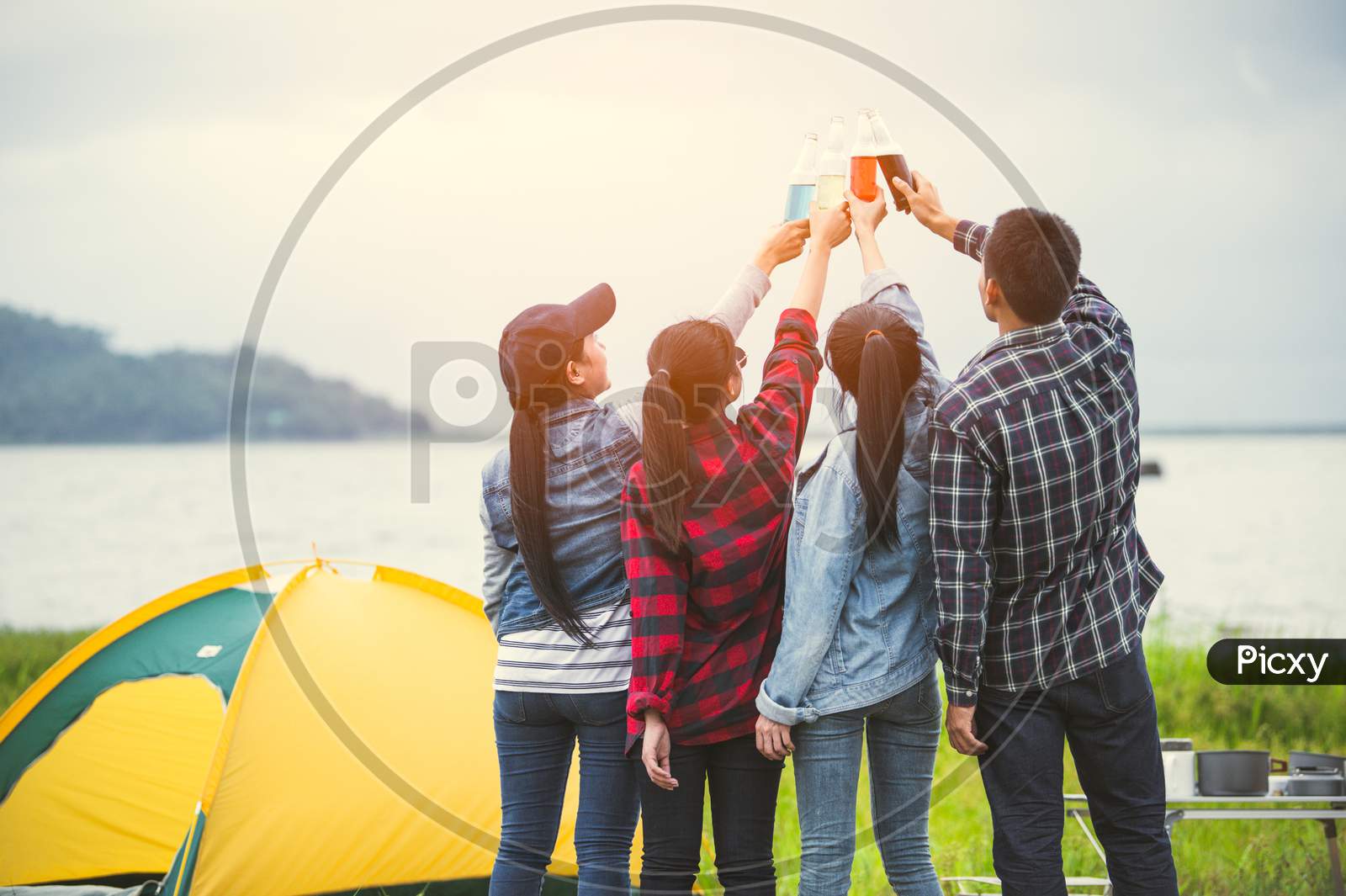 Back View Of Friendship Clinking Drinking Bottle Glass For Celebrating In Private Party With Mountain And Lake View Background. People Lifestyle And Travel On Vacation Concept. Picnic And Camping Tent