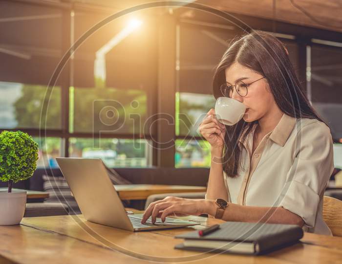 Asian Working Woman Using Laptop And Drinking Coffee In Cafe. People And Lifestyles Concept. Technology And Business Theme. Freelance And Occupation Theme. Workaholic In Overnight Concept.