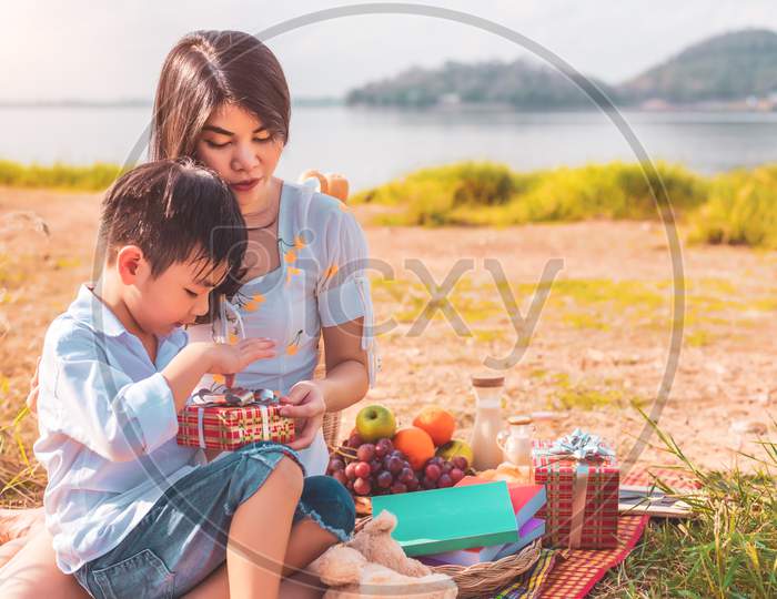Beautiful Asian Mother And Son Doing Picnic And Opening Gift Box From Surprise In Birthday Party On Meadow Near Lake And Mountain. Holiday And Vacation. People Lifestyle And Happy Family Life Concept