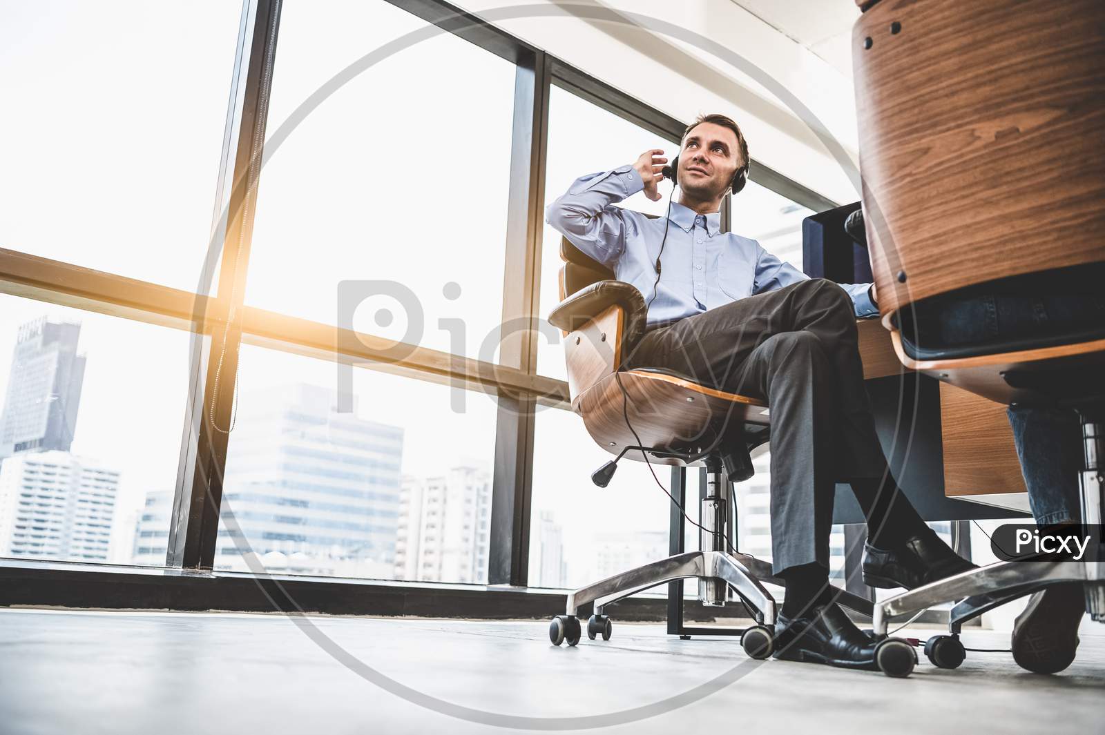 Portrait Of Happy Handsome Business Man With Headset In Modern Office With City Urban Building Background. Businessman Sitting On Chair. Caucasian Man Relaxing In Customer Care Service Call Center