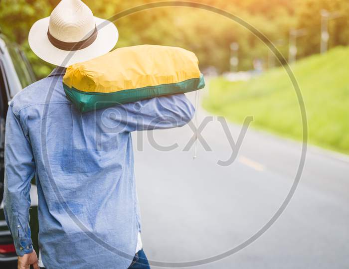 Back View Of Tourist Walking Along Road With Bag During Travel In Countryside. People Lifestyles And Vacation Concept. Man Holding And Backpack For Long Holiday Trip With Mountain Background