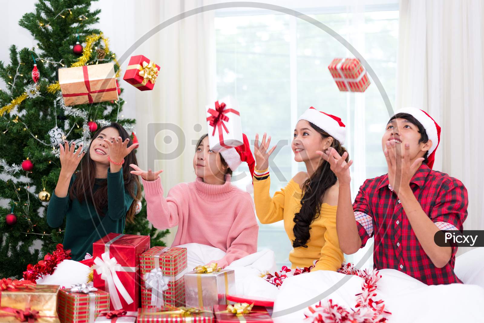 Group Of Young Asian People Throwing  Gift Box To Air With Xmas Tree Background. Holiday And Festival Concept. Christmas And New Year Event Theme. Happy People Wearing Santa Hat.