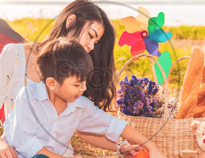 Beautiful Asian Mother And Son Doing Picnic And In Easter Summer Party On Meadow Near Lake And Mountain. Holiday And Vacation. People Lifestyle And Happy Family Life Concept. Thai Person