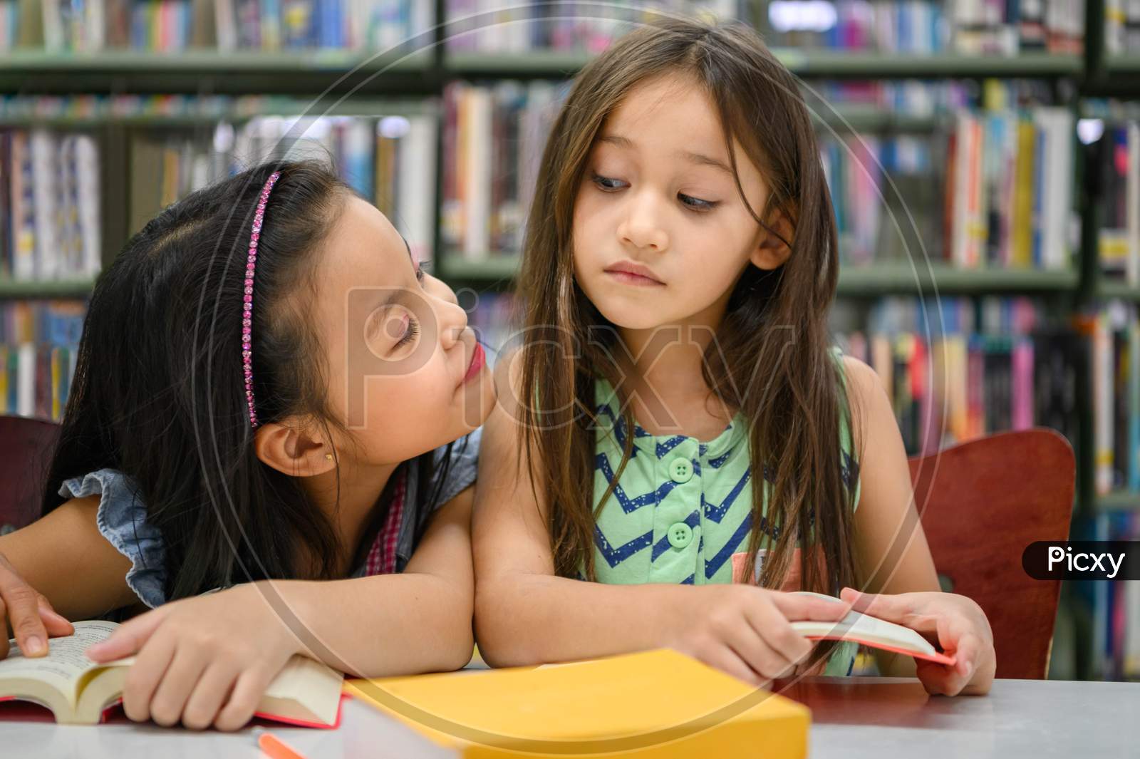 Two Cute Girls Are Jealous Of Each Other While Reading Books In Library While Teacher Teaching. People Lifestyles And Education. Young Friendship And Kids Relationship In School Concept. Face To Face