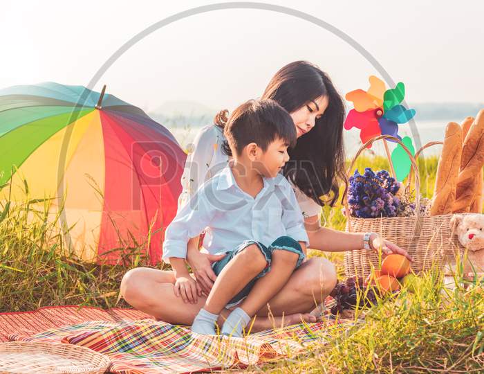 Beautiful Asian Mother And Son Doing Picnic And In Easter Summer Party On Meadow Near Lake And Mountain. Holiday And Vacation. People Lifestyle And Happy Family Life Concept. Thai Person