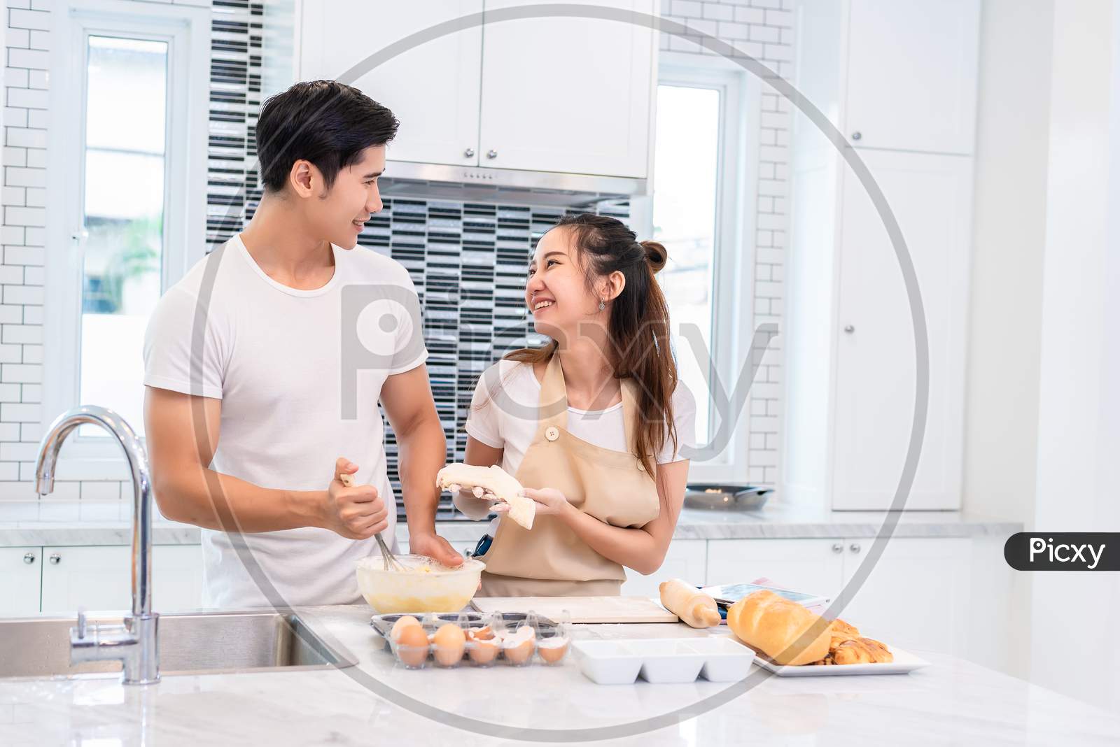 Asian Couples Cooking And Baking Cake Together In Kitchen Room. Man And Woman Looking To Each Other At Home. Love And Happiness Concept Sweet Honeymoon And Valentine Day Theme