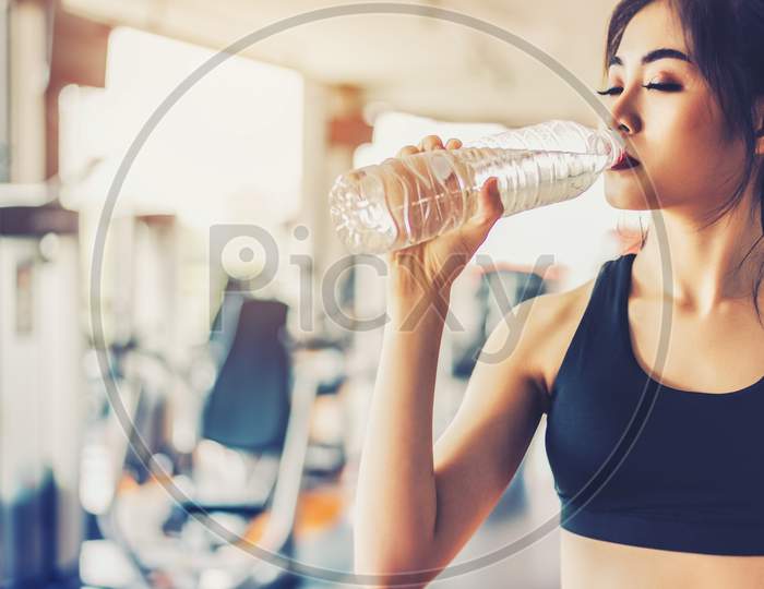 Asian Woman Drinking Pure Drinking Water For Freshness After Workout Or Exercise Training In Fitness Gym With Fitness Equipment Background. Relax And Rest Concept. Strength And Fitness Training Theme.
