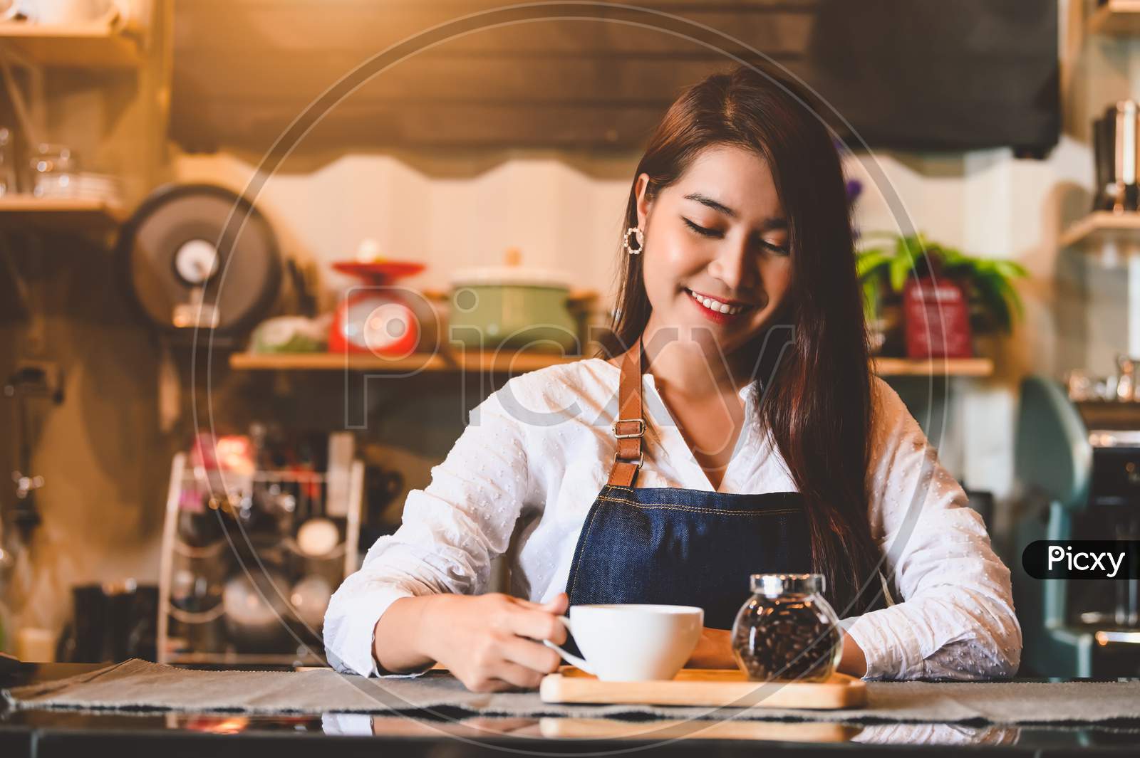 Asian Female Barista Making Cup Of Coffee. Young Woman Holding White Coffee Cup While Standing Behind Cafe Counter Bar In Restaurant Background. People Lifestyles And Business Occupation Concept.