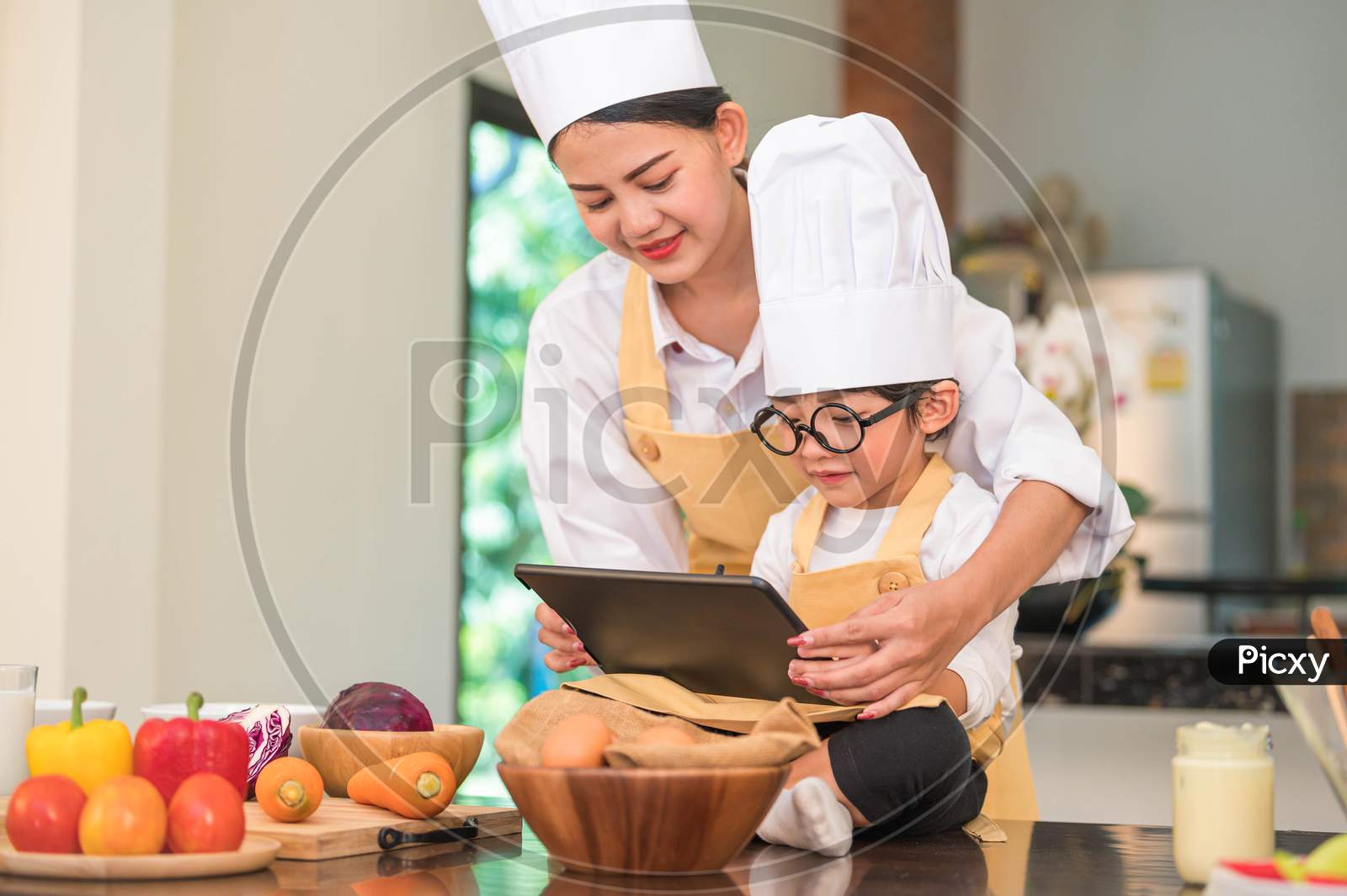 Beautiful Asian Woman And Cute Little Boy With Eyeglasses Prepare To Cooking In Kitchen At Home With Tablet. People Lifestyles And Family. Homemade Food And Ingredients Concept. Ketogenic Salad Making