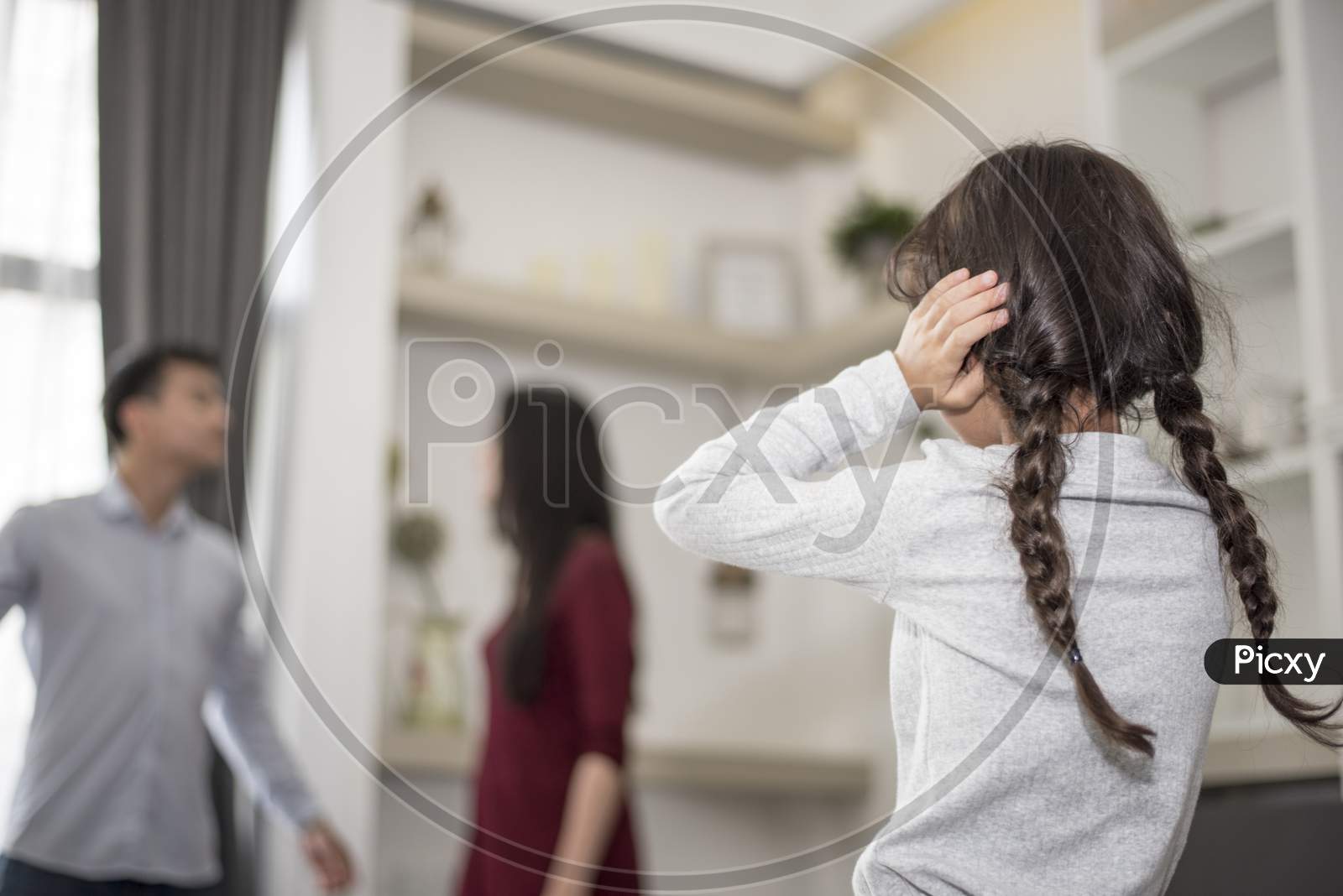 In Back View, Little Girl Puts Her Hands On Her Ears Because She Does Not Want To Hear Her Dad And Mom Quarrel. Close Ears, Family Dramatic Scene, Parrents Issues, Social And Parents Problem Concept