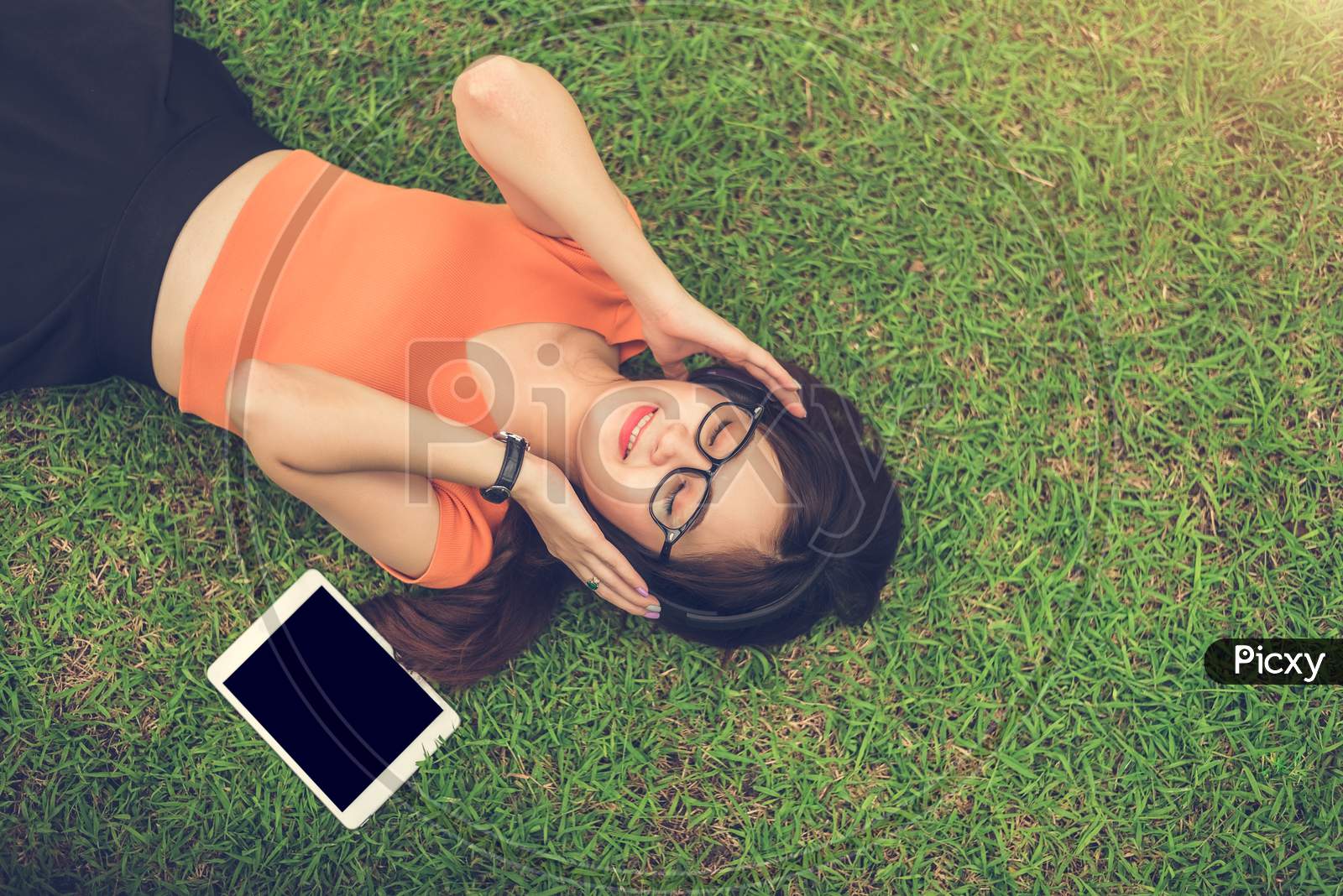 Asian Woman Listening Music With Blank Tablet Black Isolated Screen On Grass In Park. People Lifestyle And Entertainment Concept.