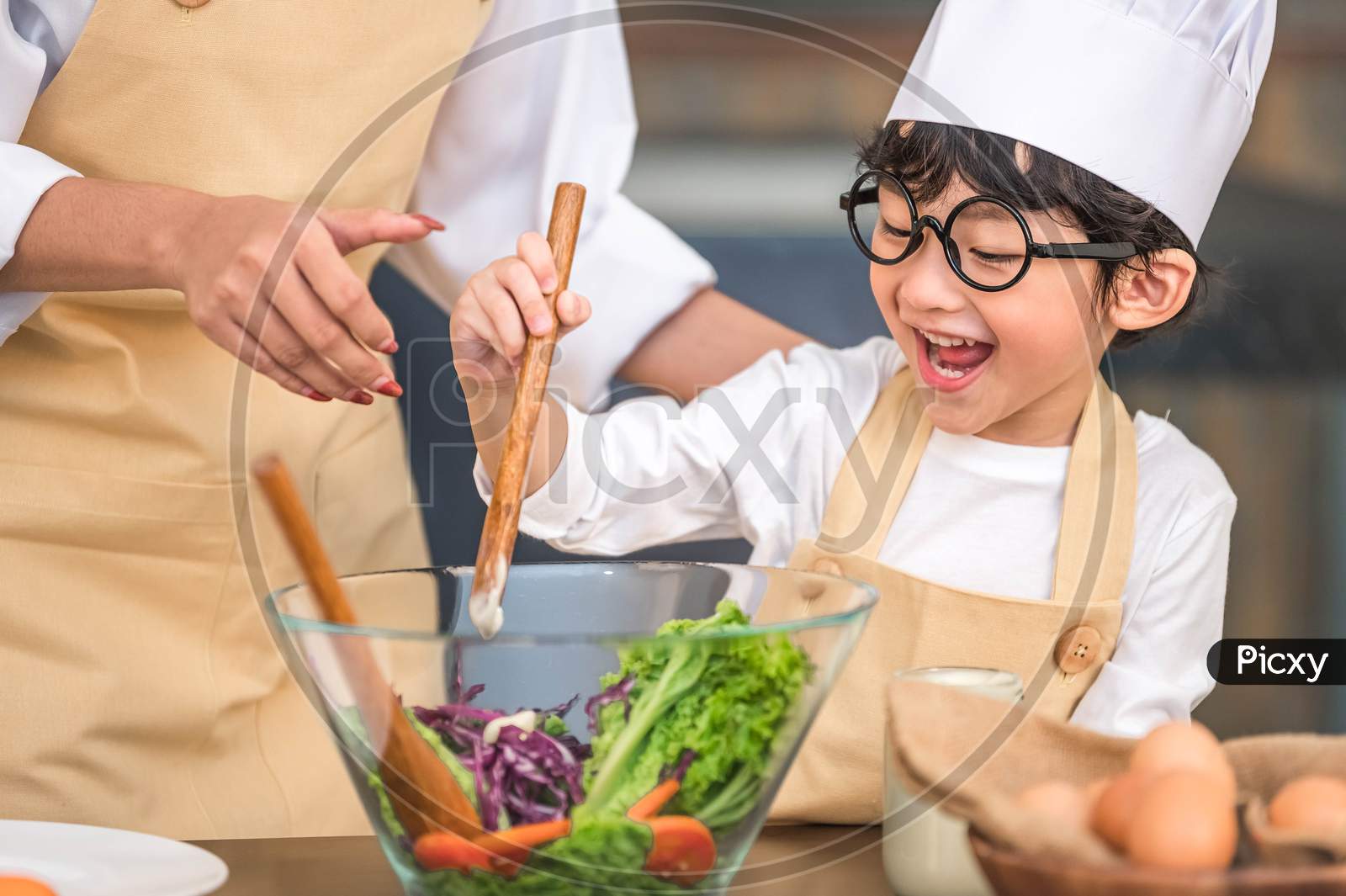 Cute Little Asian Happy Boy Interested In Cooking With Mother Funny In Home Kitchen. People Lifestyles And Family. Homemade Food And Ingredients Concept. Two Thai People Making Ketogenic Diet Salad