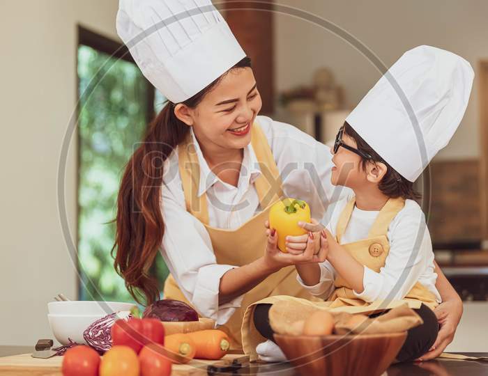 Happy Asian Family In Kitchen. Mother And Son In Chef Hat Preparing Food In Home Kitchen Together. People Lifestyle And Family. Homemade Food And Ingredients Concept. Two Thai People In Teaching Class