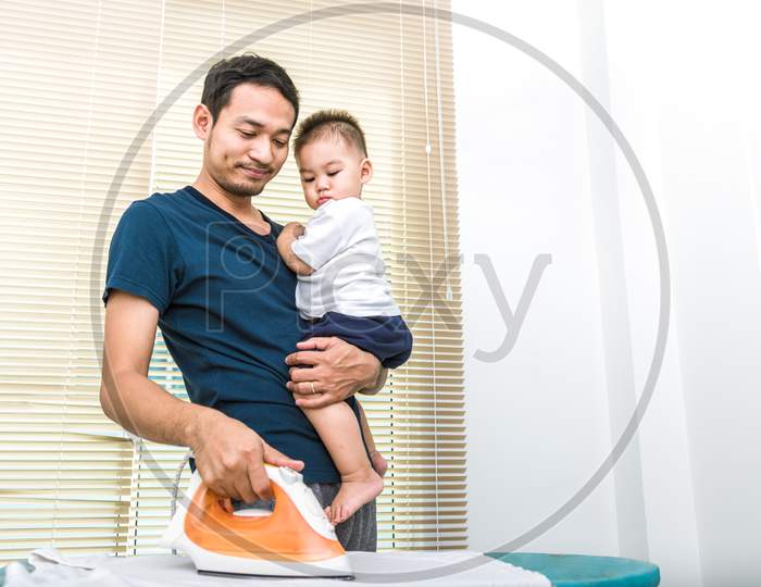 Single Dad Is Ironing While Carrying His Son. People And Lifestyles Concept.