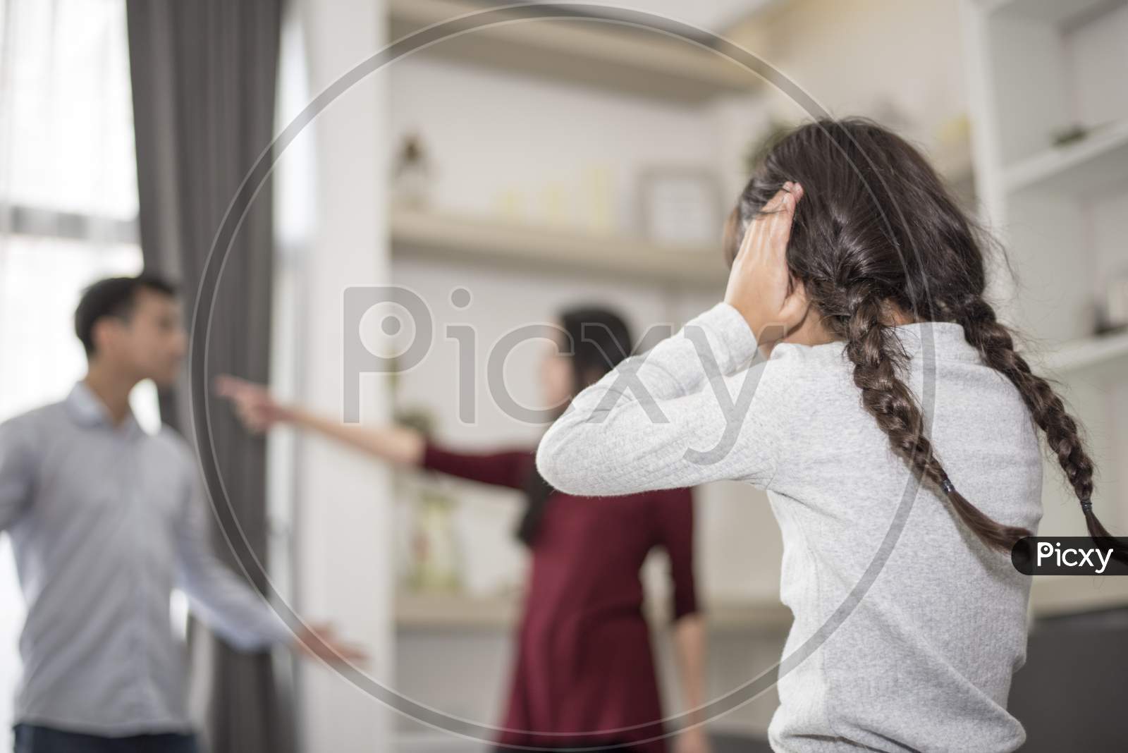 In Back View, Little Girl Puts Her Hands On Her Ears Because She Does Not Want To Hear Her Dad And Mom Quarrel. Close Ears, Family Dramatic Scene, Parrents Issues, Social And Parents Problem Concept
