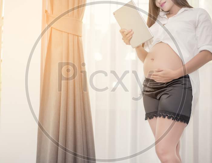 Pregnant Woman Standing And Using The Tablet With Touch Her Bellow In Her House, Family And Health Care Concept