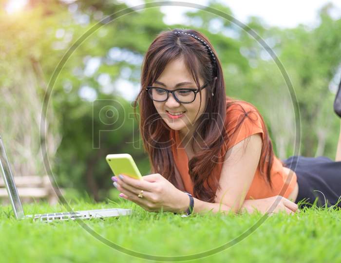 Happy Asian Woman Using Smartphone When Relaxing In Park. People And Lifestyle Concept. Technology And Relaxation Theme. Outdoors Leisure And Activity Theme. Laptop Computer Element
