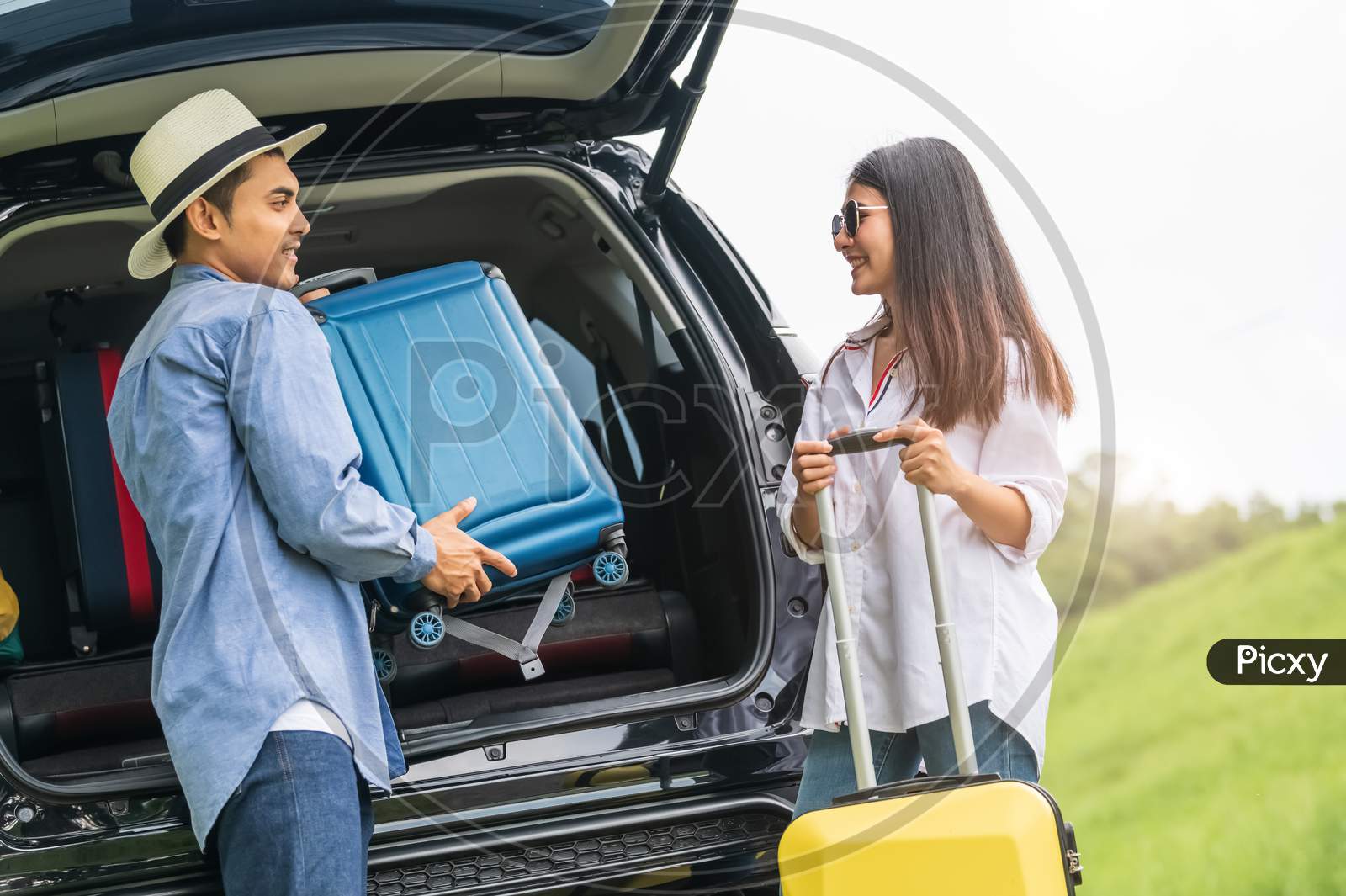 Asian Man Helping Woman To Lifting Suitcase From Car During Travel In Long Weekend. Couple Have Road Trip In Vacation With Yellow Luggage. People Lifestyle And Transportation Concept. Nice Guy Theme