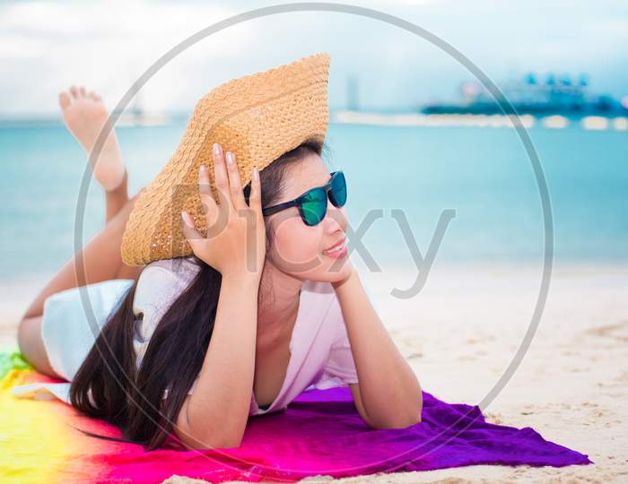 Asian Woman In Casual And Straw Hat Lying On Tropical Beach With Sea Background. Relax And Lifestyle Concept. Holiday And Vacation Concept. Single Woman And Happiness Theme.