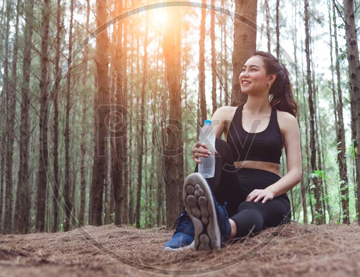 Beauty Asian Sport Woman Resting And Holding Drinking Water Bottle And Relaxing In Middle Of Forest After Tired From Jogging. Girl Sitting And Looking Attraction View. Workout Concept. Lifestyle Theme