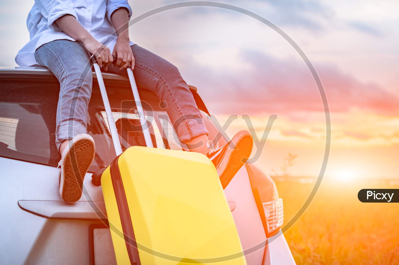 Closeup Of Happy Asian Woman On Top Of Car With Luggage Bag. Girl Sitting On Roof And Looking Sunset Before Evening. People Lifestyle In Long Vacation Trip Concept. Nature And Transportation Vehicle