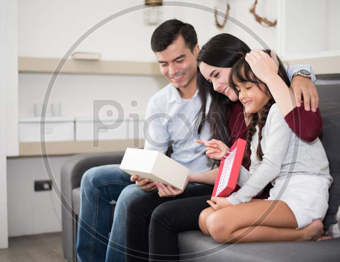 Father And Mother Surprise Their Daughter By Gift Or New Toy. Parents And Children Are Happy Together In Home On Sofa. Family And Happiness Concept.