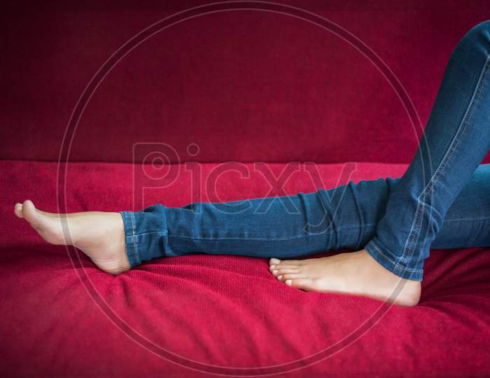 Woman Legs Wear Jeans On Red Sofa At Home, Relax And Beauty Concept