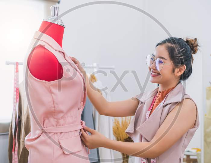 Happy Asian Female Fashion Designer Girl Making Fit On The Formal Dress Uniform Clothes On Mannequin Model. Fashion Designer Stylish Showroom. Sewing And Tailor Concept. Creative Dressmaker Stylist.