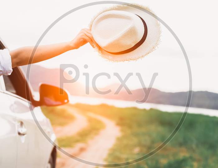Happy Woman Hand Holding Hat Outside Open Window Car With Meadow And Mountain Lake Background. People Lifestyle Relaxing As Traveler On Road Trip In Holiday Vacation. Transportation And Travel Concept