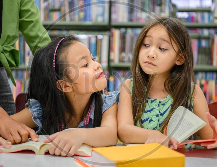Two Cute Girls Are Jealous Of Each Other While Reading Books In Library While Teacher Teaching. People Lifestyles And Education. Young Friendship And Kids Relationship In School Concept. Daycare Theme