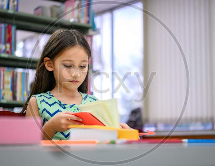 Cute Little Girl Reading Books In Library Attentively At School. Girl Concentrating To Learning By Herself Outside Classroom. Education And People Lifestyles. Academic And Kids Duty Concept.