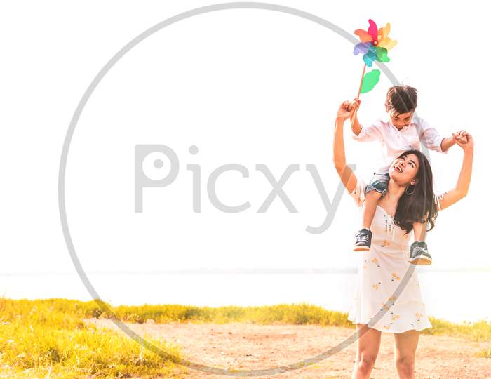 Little Asian Boy Riding Back On His Super Power Mom In Meadow Near Lake. Mother And Son Playing Together. Celebrating In Mother Day And Appreciating Concept. Summer People And Lifestyle Theme.