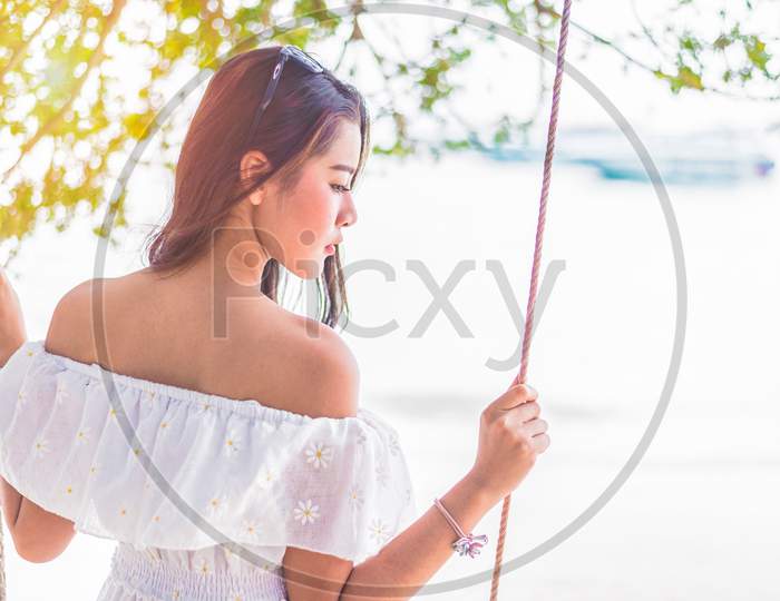 Asian Woman On White Dress Sitting On Swing At Beach. People And Nature Concept. Sad Love And Missing Someone Concept. Lonely And Heart Broken Theme.