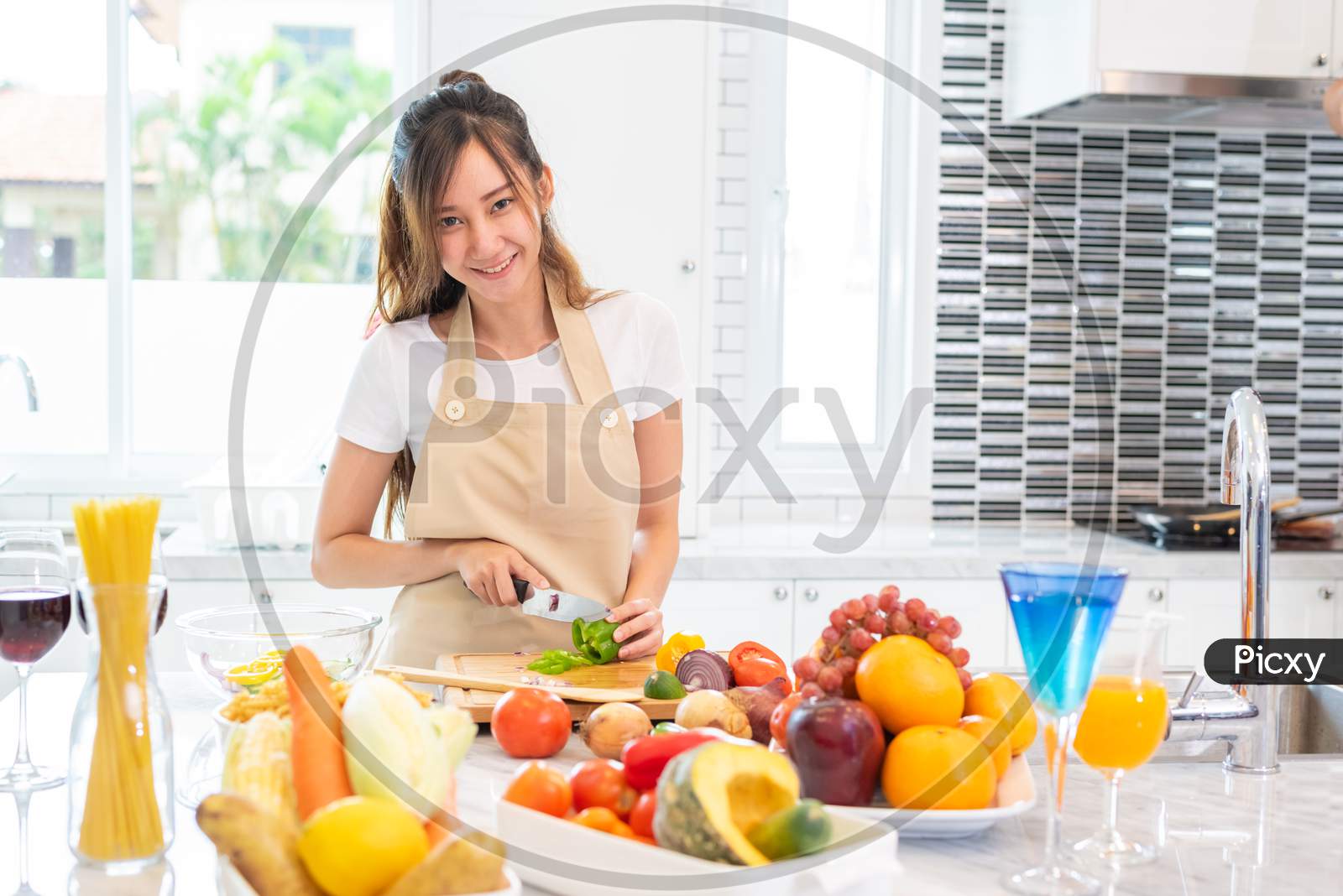 Asian Beauty Woman Cooking And Slicing Vegetable In Kitchen Room With Full Of Food And Fruit On Table For Party. Holiday And Happiness Concept. People And Lifestyles Concept. Girl Looking At Camera