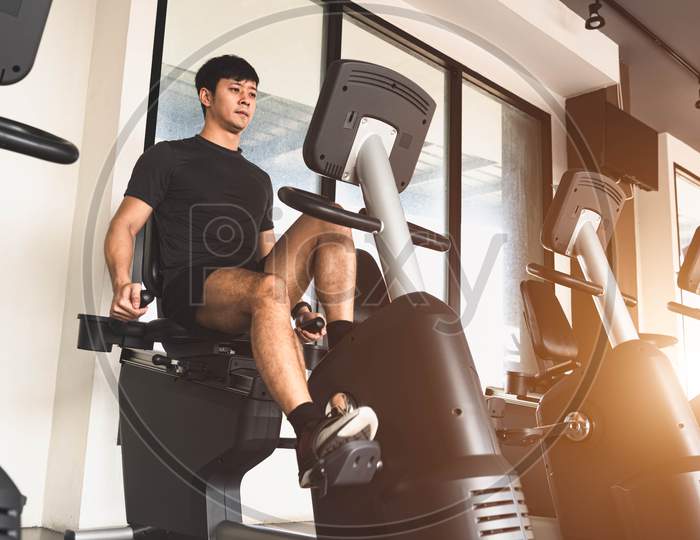 Asian Young Sport Man Riding Stationary Bicycle In Fitness Gym. Man Working Out On Spinning Bikes In Gym. People Lifestyles And Health Club Concept. Bodybuilding Theme