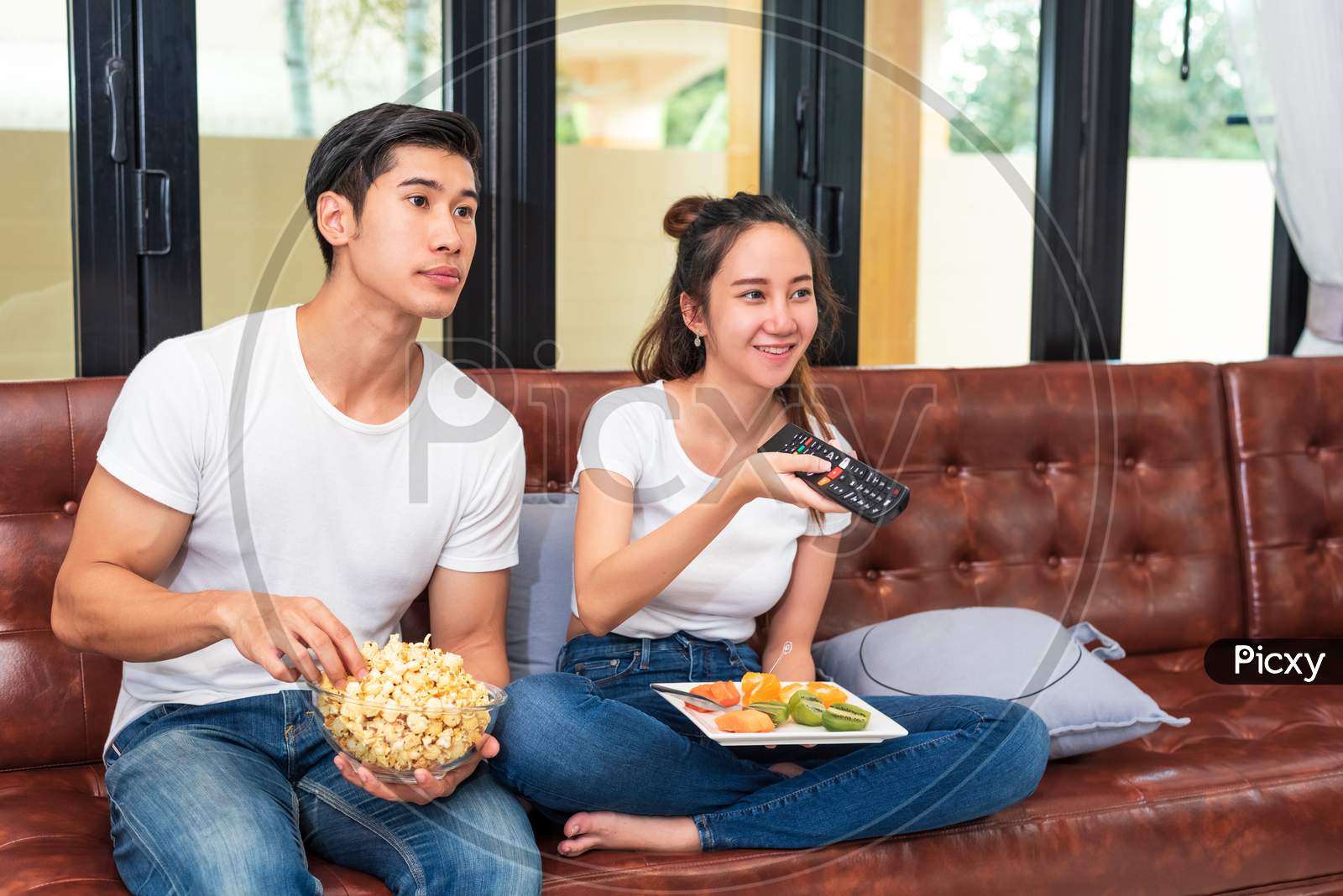 Asian Couples Watching Television Together On Sofa In Their Home. People And Lifestyles Concept. Vacation And Holiday Concept. Honeymoon And Pre Wedding Theme. Happy Family Activity In Valentines Day