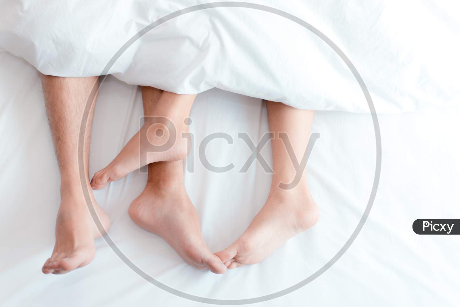 Closeup Feet Of Couple On The Bed. Man And Woman Lovers Make Love Under The Blanket Or Bed Sheet. Sex On Vacation Theme. Valentine And Honeymoon Concept.