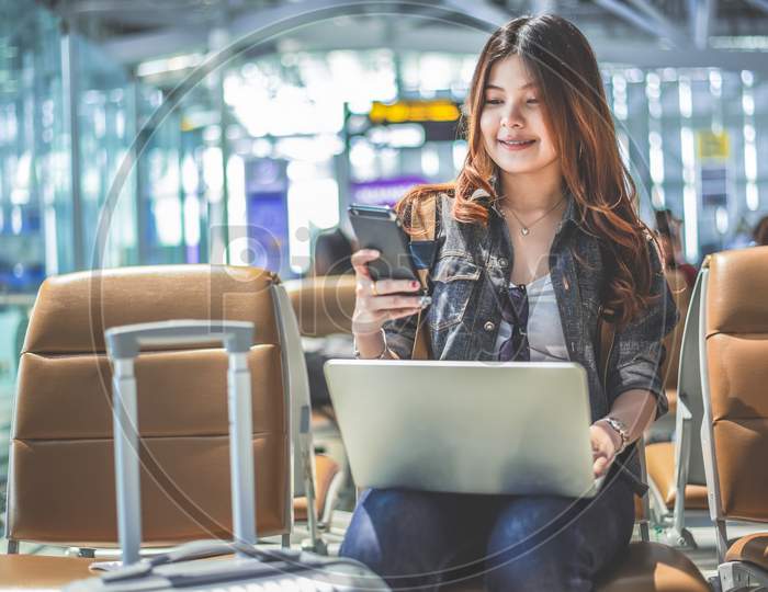 Young Asian Female Passenger Using Laptop And Smart Phone While Sitting On Seat In Terminal Hall And Waiting For Flight In Airport . People Lifestyles And Happy Woman. Technology And Travel Concept.