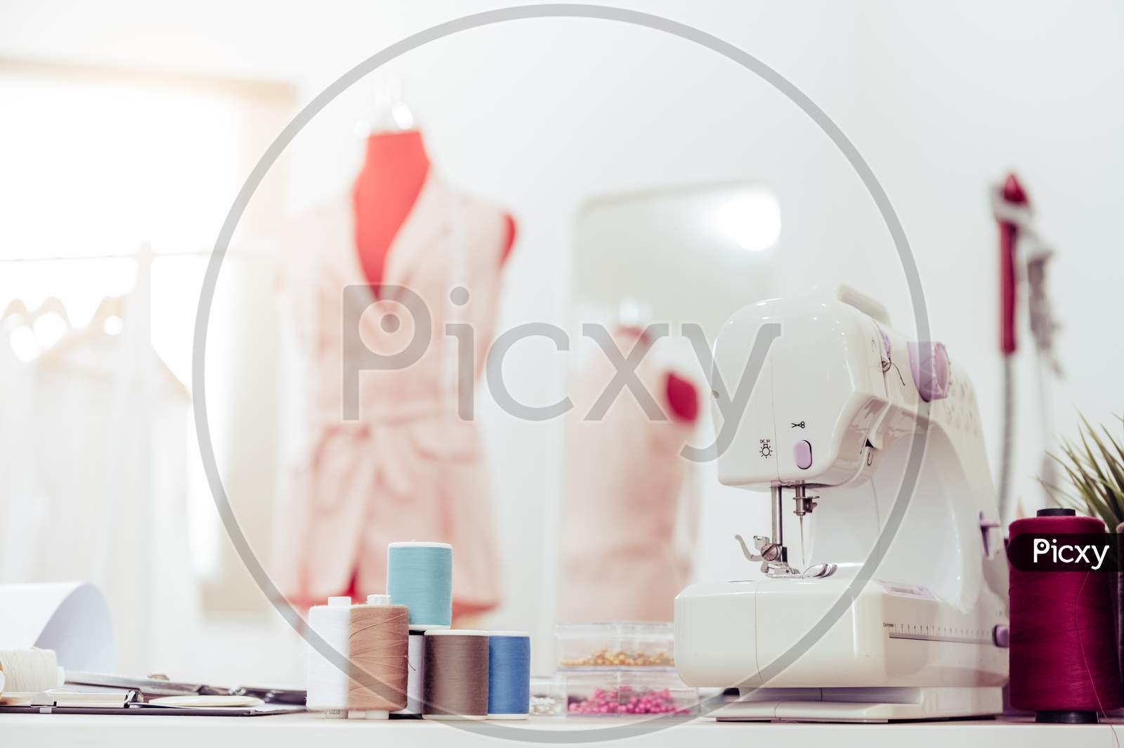 Closeup Of Sewing Machine With Fashion Designer Showroom Studio Workshop Background With New Collection Of Pink Pastel Female Clothes Design. Tailor And Sewing Concept. Dressmaker Modern Room Interior
