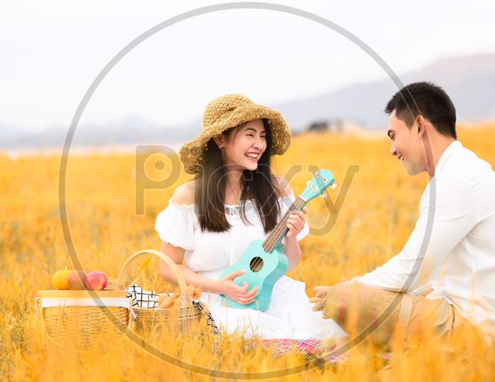 Two Asian Young Couples In Autumn Meadow Field Doing Picnic In Honeymoon Trip In White Clothes, Ukulele Guitar And Fruits Basket. People Lifestyle And Wedding Concept. Nature And Travel Day Concept.