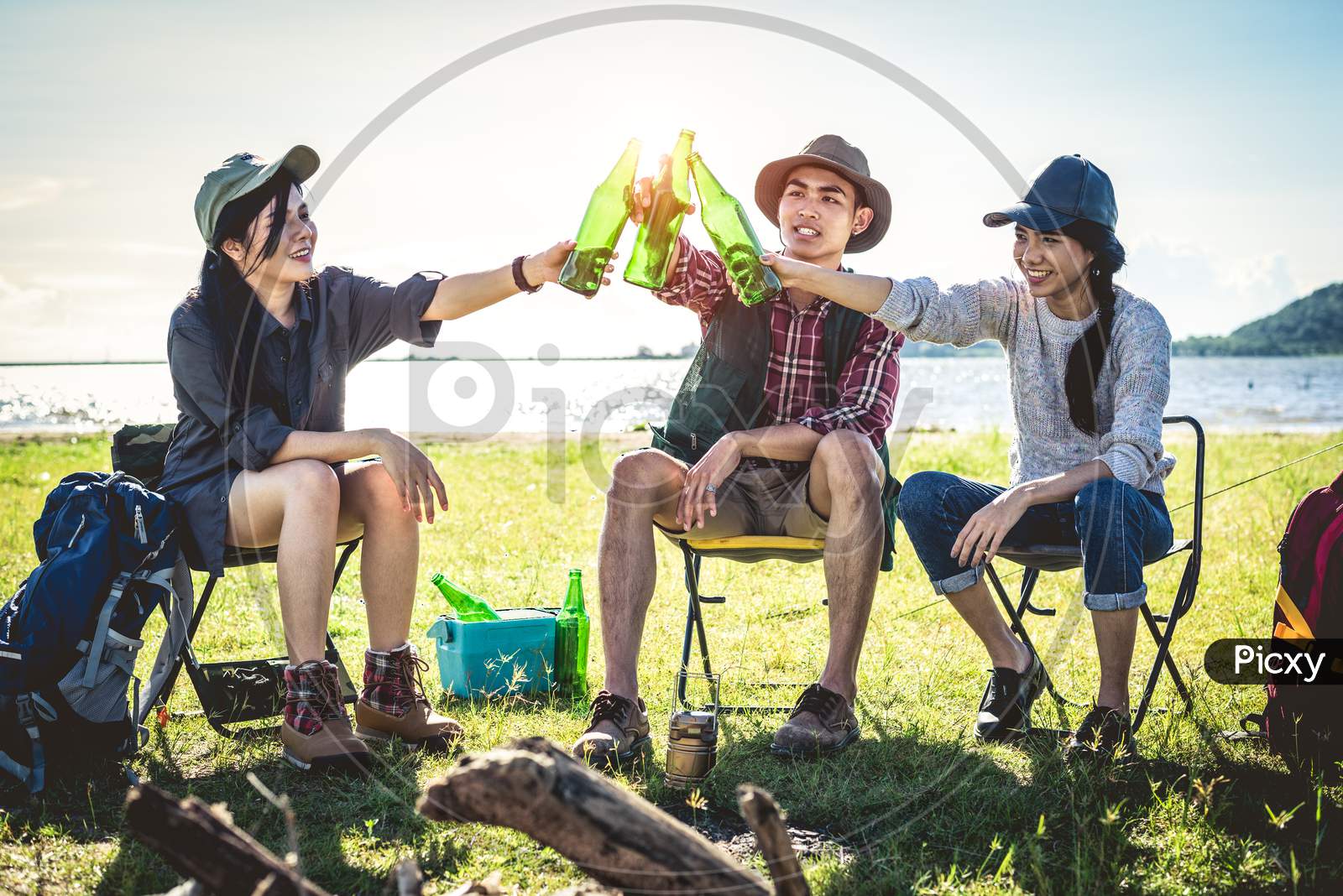Group Of Young Asian Friends Enjoy Picnic And Party At Lake With Camping Backpack And Chair. Young People Toasting And Cheering Bottles Of Beer. People And Lifestyles Concept. Outdoor Background Theme