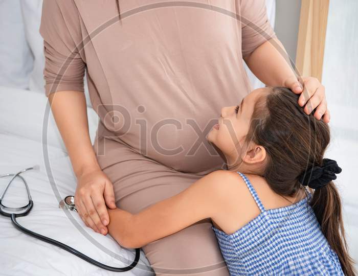 Cute Daughter Hugging Mother And Listening To Voice Of Baby In Mother Belly For Checking And Looking At Mom Face. Pregnant And Parents. Health And Medical. People Lifestyles And Family Love Concept.