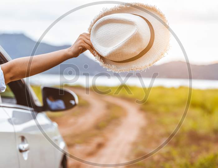 Happy Woman Hand Holding Hat Outside Open Window Car With Meadow And Mountain Lake Background. People Lifestyle Relaxing As Traveler On Road Trip In Holiday Vacation. Transportation And Travel Concept